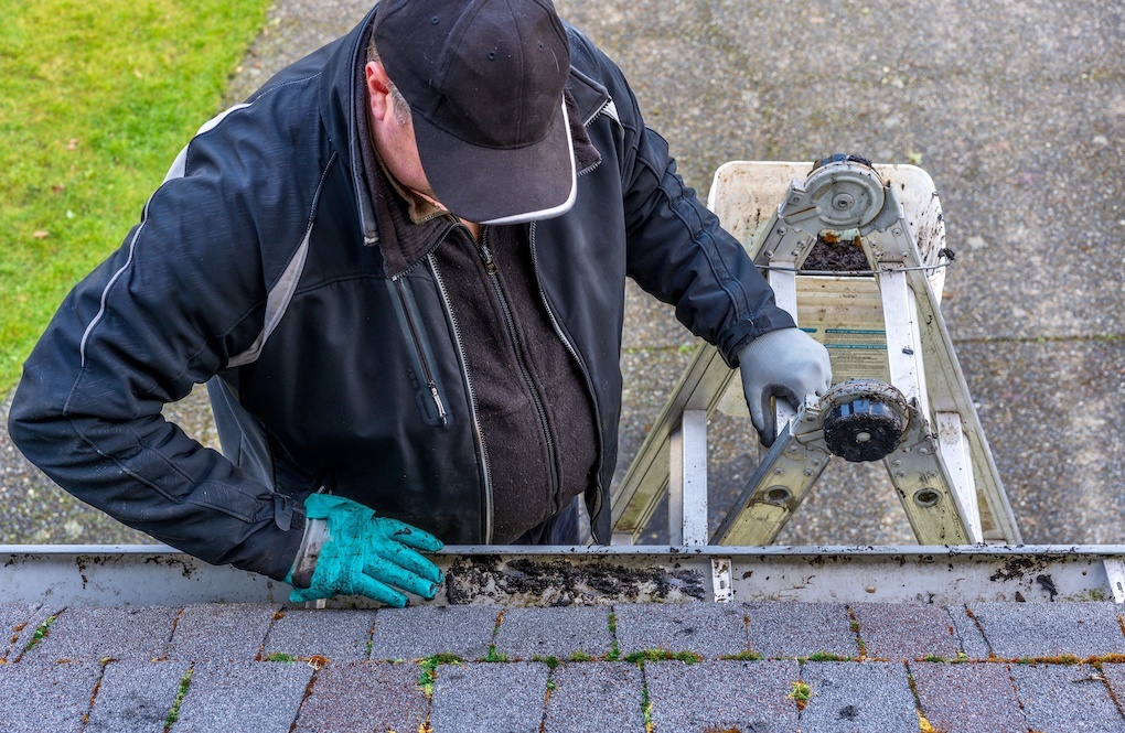 Man on top of ladder cleaning dirt from clogged roof gutters to determine roof repair cost
